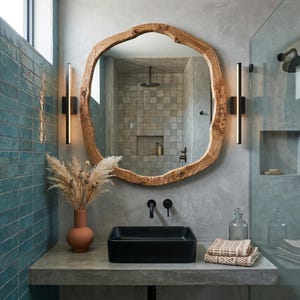 May include: A bathroom vanity featuring a large, organic-shaped wooden mirror. A black rectangular sink is set on a concrete countertop. Decorative items include a terracotta vase with dried flowers, two black sconces, and a glass bottle.
