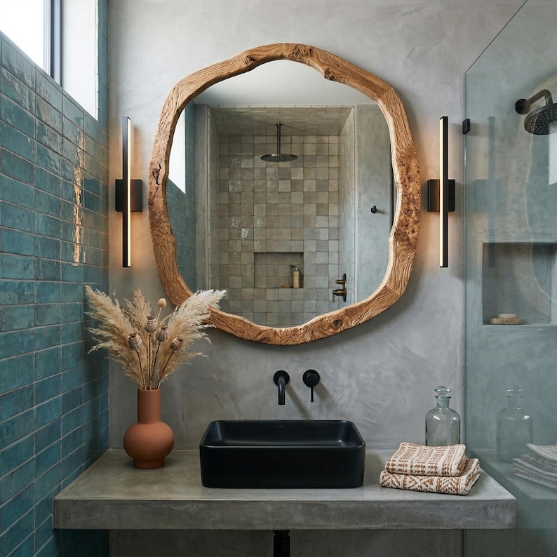 May include: A bathroom vanity featuring a large, organic-shaped wooden mirror. A black rectangular sink is set on a concrete countertop. Decorative items include a terracotta vase with dried flowers, two black sconces, and a glass bottle.