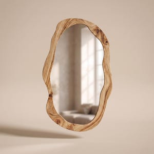 May include: An oval-shaped mirror with a natural wood frame. The frame has a unique, organic shape with a light brown color and visible wood grain. The mirror reflects a room with a window and furniture.