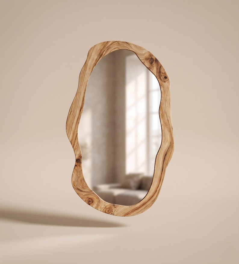 May include: An oval-shaped mirror with a natural wood frame. The frame has a unique, organic shape with a light brown color and visible wood grain. The mirror reflects a room with a window and furniture.