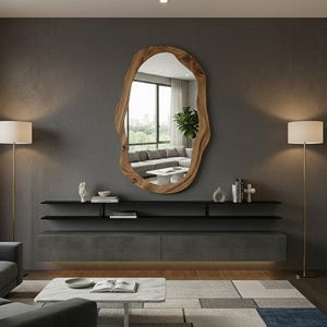 May include: A large, irregularly shaped wooden mirror hangs on a gray wall. Below the mirror is a dark gray floating shelf and a matching cabinet. Two gold floor lamps flank the mirror, illuminating the room. The mirror reflects a living room with a gray sofa.