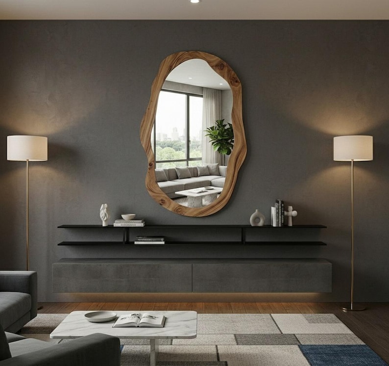 May include: A large, irregularly shaped wooden mirror hangs on a gray wall. Below the mirror is a dark gray floating shelf and a matching cabinet. Two gold floor lamps flank the mirror, illuminating the room. The mirror reflects a living room with a gray sofa.