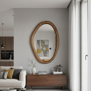 May include: A large, organically shaped wooden mirror with a warm brown finish hangs on a gray wall. The mirror reflects a room with a sofa, a wooden cabinet, and a piece of abstract art. The room is bathed in natural light from a nearby window.