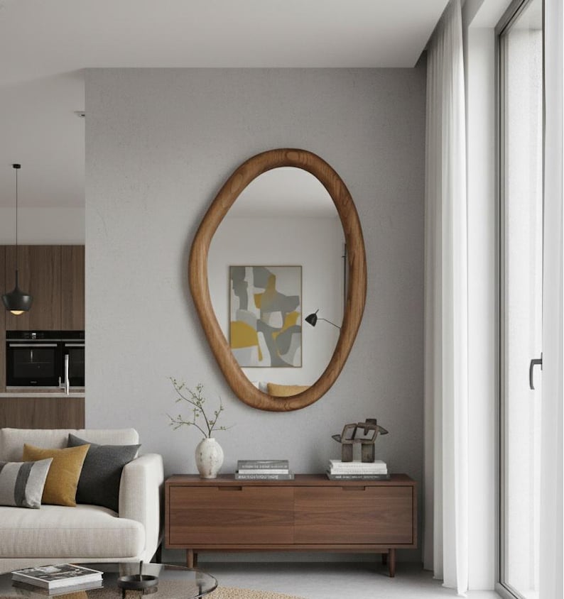 May include: A large, organically shaped wooden mirror with a warm brown finish hangs on a gray wall. The mirror reflects a room with a sofa, a wooden cabinet, and a piece of abstract art. The room is bathed in natural light from a nearby window.