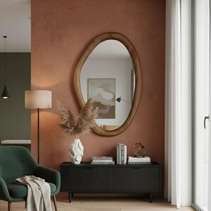 May include: An oval-shaped wooden mirror with a natural wood grain finish hangs on a textured, reddish-brown wall. Below the mirror is a black cabinet with books, a decorative object, and a white vase with dried plants. A green armchair and floor lamp are in the foreground.