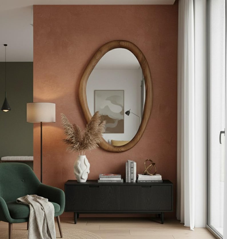 May include: An oval-shaped wooden mirror with a natural wood grain finish hangs on a textured, reddish-brown wall. Below the mirror is a black cabinet with books, a decorative object, and a white vase with dried plants. A green armchair and floor lamp are in the foreground.