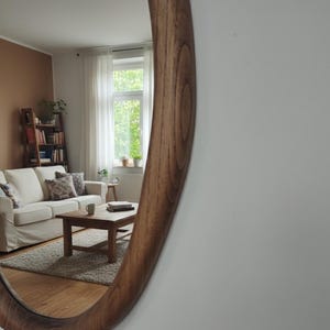 May include: A large, oval-shaped wooden mirror with a warm, brown finish. The mirror reflects a living room scene with a white sofa, wooden coffee table, and a window with natural light. The mirror's frame has a smooth, curved design.