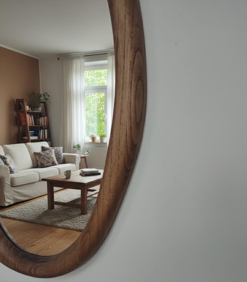 May include: A large, oval-shaped wooden mirror with a warm, brown finish. The mirror reflects a living room scene with a white sofa, wooden coffee table, and a window with natural light. The mirror's frame has a smooth, curved design.