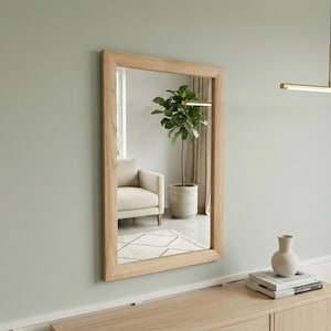 Peut inclure: Un miroir rectangulaire encadré de bois est accroché à un mur vert pâle. Le miroir reflète une pièce avec un fauteuil beige, une plante en pot et un tapis. Un petit vase clair est posé sur un meuble en bois.