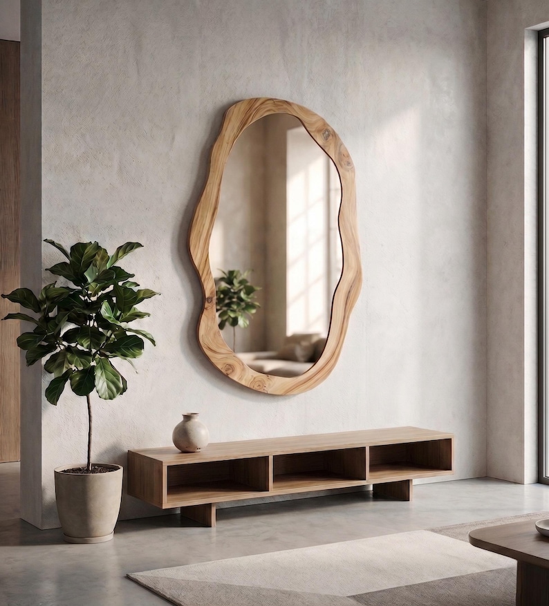 May include: A large, organically shaped wooden mirror hangs on a neutral-toned wall. Below, a low, rectangular wooden console with open compartments and a small vase sits on the floor. A potted plant is to the left.