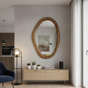 May include: A large, organically shaped wooden mirror hangs on a textured wall. Below, a light wood cabinet with metal legs holds small potted plants and a black speaker. A modern floor lamp with a globe light and a blue armchair complete the room's decor.