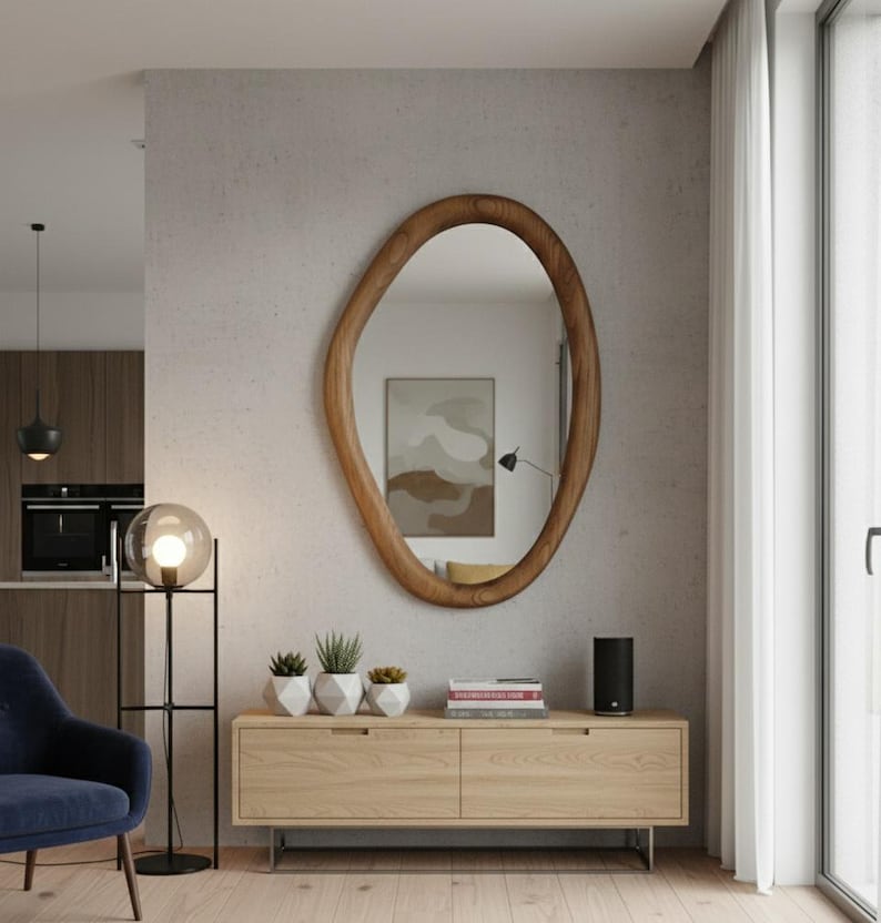May include: A large, organically shaped wooden mirror hangs on a textured wall. Below, a light wood cabinet with metal legs holds small potted plants and a black speaker. A modern floor lamp with a globe light and a blue armchair complete the room's decor.