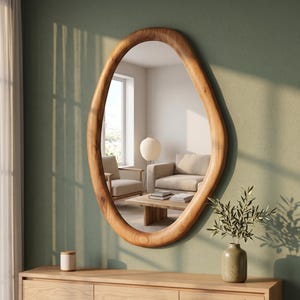 May include: An oval wall mirror with a thick, smooth, natural light brown wooden frame. The mirror reflects a room with a beige armchair, a wooden coffee table, and a paper floor lamp. The frame has a visible wood grain texture throughout.