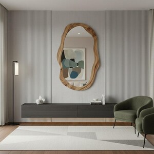 May include: A modern living room featuring a large, organically shaped wooden mirror. Below the mirror is a dark gray cabinet, with two green armchairs and a geometric patterned rug. A black floor lamp stands to the left.