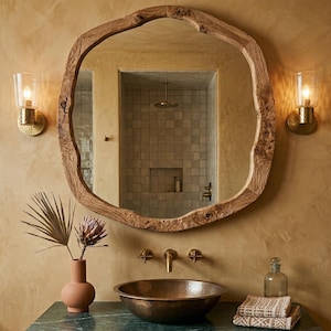May include: A rustic bathroom scene featuring a large, irregularly shaped wooden mirror. The mirror reflects a shower area with square tiles. Below, a copper sink sits on a green countertop, complemented by a vase with dried flowers and two sconces.