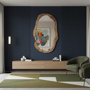 May include: A modern living room featuring a large, organic-shaped wooden mirror. Below the mirror is a light wood floating cabinet. Two green armchairs sit on a geometric patterned rug. The wall is a dark blue with vertical paneling.