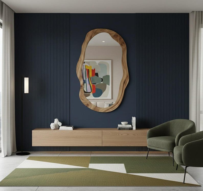 May include: A modern living room featuring a large, organic-shaped wooden mirror. Below the mirror is a light wood floating cabinet. Two green armchairs sit on a geometric patterned rug. The wall is a dark blue with vertical paneling.
