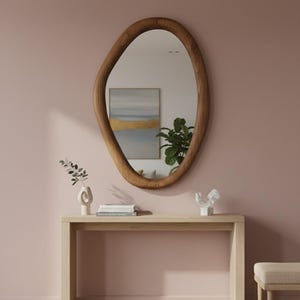 May include: An oval-shaped wooden mirror with a natural wood finish hangs on a pink wall. Below the mirror is a light wood console table with decorative items, including a vase, books, and a sculpture. The room has a minimalist, modern aesthetic.