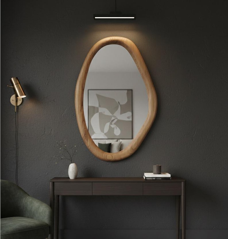 May include: An oval-shaped wooden framed mirror hangs on a dark gray wall. A small dark brown console table sits below the mirror, with a vase, books, and a candle on top. A gold wall sconce and a black light fixture are also visible.