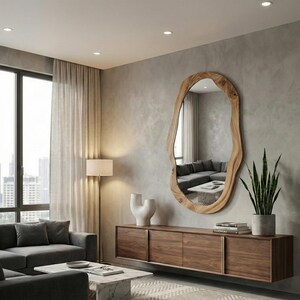 May include: A large, irregularly shaped wooden mirror hangs on a gray wall. Below, a dark wood cabinet with sliding doors holds books and a potted plant. A gray sofa and marble coffee table complete the modern living room.