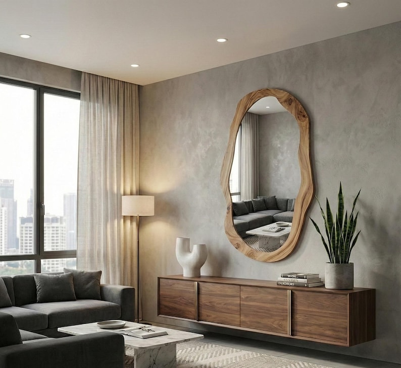 May include: A large, irregularly shaped wooden mirror hangs on a gray wall. Below, a dark wood cabinet with sliding doors holds books and a potted plant. A gray sofa and marble coffee table complete the modern living room.