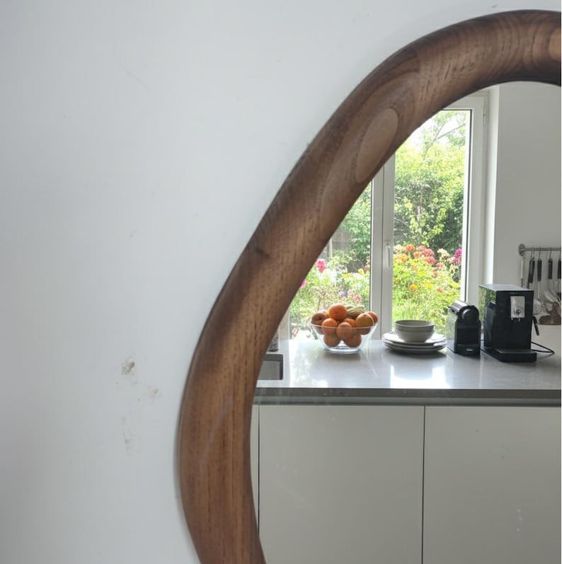 May include: A large, round mirror with a dark wood frame. The mirror reflects a kitchen scene with a window overlooking a garden, a bowl of oranges, and a coffee machine.