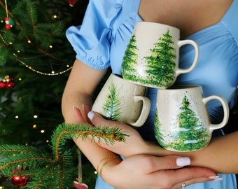 May include: Three ceramic mugs with hand-painted Christmas tree designs. The mugs are stacked on top of each other and are being held by a person. The mugs are white with green and brown paint.