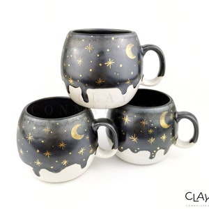 May include: Three black ceramic mugs with white rims and gold painted stars and crescent moons. The mugs are stacked on top of each other.