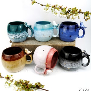 May include: Six ceramic mugs with a bird handle. The mugs are in various colours, including teal, blue, pink, brown, black, and a light blue. Each mug has a speckled glaze and gold accents.
