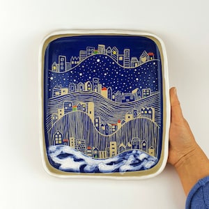 Puede incluir: Una bandeja cuadrada de servir de cerámica blanca con fondo azul y una ilustración dorada y blanca de una ciudad con casas y estrellas.