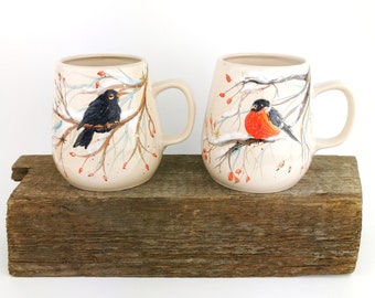 May include: Two ceramic mugs with winter bird designs. One mug features a blackbird on a branch with red berries, while the other mug features a robin on a branch with red berries. Both mugs have a cream-colored background with a snowy, wintery scene.