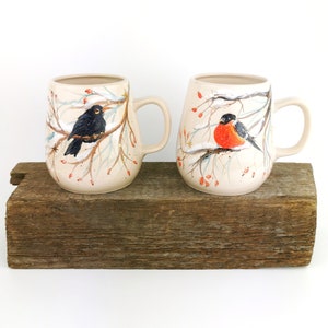 May include: Two ceramic mugs with winter bird designs. One mug features a blackbird on a branch with red berries, while the other mug features a robin on a branch with red berries. Both mugs have a cream-colored background with a snowy, wintery scene.