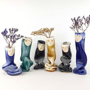 Puede incluir: Seis jarrones de cerámica con caras únicas y diferentes colores, incluyendo azul, verde, negro, dorado y blanco. Los jarrones están dispuestos en una fila sobre una superficie blanca. Cada jarrón tiene una pequeña abertura en la parte superior para flores u otros elementos decorativos.