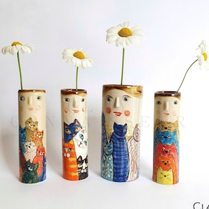 May include: Four ceramic vases with a whimsical design featuring a woman's face and a cluster of colourful cats. Each vase is decorated with a different pattern of cats, including orange, blue, and yellow. The vases are arranged in a row on a white surface, with a single daisy in each vase.