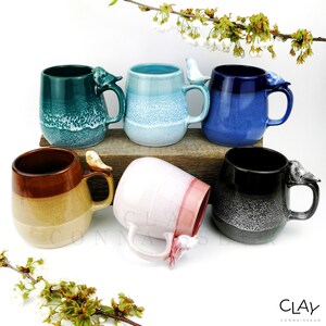 May include: Six ceramic mugs with bird handles. The mugs are in various colours, including teal, blue, pink, brown, and black. Each mug has a different glaze finish. The mugs are arranged on a wooden surface.
