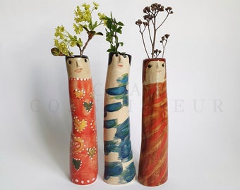 Puede incluir: Tres jarrones de cerámica con caras pintadas y diseños abstractos. Los jarrones están decorados con pintura roja, naranja, azul y blanca. Los jarrones están llenos de flores secas y vegetación.