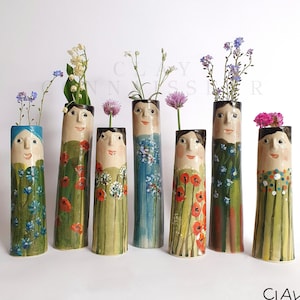 Könnte beinhalten: Sieben Keramikvasen mit bemalten Gesichtern und Blumendesigns. Die Vasen sind grün, blau und braun mit bunten Blumen und Mustern.