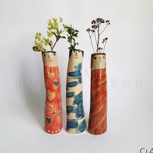 Puede incluir: Tres jarrones de cerámica con caras pintadas y diseños abstractos. Los jarrones están decorados con pintura roja, naranja, azul y blanca. Los jarrones están llenos de flores secas y vegetación.