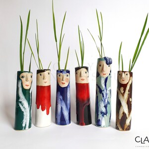 Puede incluir: Seis jarrones de cerámica con caras pintadas y esmaltes coloridos. Cada jarrón tiene un diseño y un color diferente, incluyendo verde, rojo, azul, negro y marrón. Los jarrones están llenos de plantas verdes.