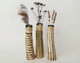 May include: Three ceramic vases with faces and striped patterns. Each vase is holding dried flowers.