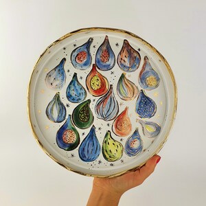 May include: A white ceramic plate with a gold rim features a colourful design of figs. The figs are painted in shades of blue, green, red, and yellow. The plate is decorated with gold stars.