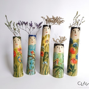 Puede incluir: Cinco jarrones de cerámica con caras pintadas y diseños florales. Cada jarrón es de un color diferente y presenta un patrón floral único, que incluye amapolas, pájaros y hojas.