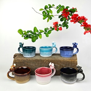 May include: Six ceramic mugs with a small elephant figurine on the handle. The mugs are in various colours, including teal, blue, pink, brown, and black. Each mug has a white glaze on the bottom half.