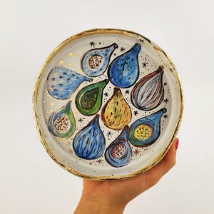 Op de afbeelding: Een wit keramiek bord met een gouden rand heeft een handgeschilderd ontwerp van vijgen. De vijgen zijn geschilderd in tinten blauw, groen, geel en bruin. Het bord is versierd met gouden sterren.
