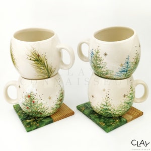 May include: Two sets of stacked ceramic mugs with a winter forest design. Each mug has a green, blue, and gold forest scene with red berries. The mugs are sitting on green and wood coasters.