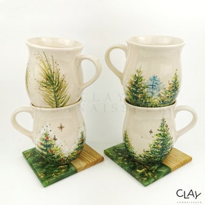 May include: Four ceramic mugs with a winter forest design. The mugs are stacked in pairs and are sitting on green and wood coasters. The mugs are white with green, blue, and gold accents.