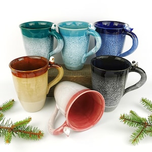 May include: A collection of six ceramic mugs in various colors, including teal, blue, and black. Each mug features a unique speckled glaze and a handle adorned with a small bird figurine. The mugs are displayed on a wooden surface with evergreen sprigs.
