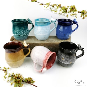 May include: Six ceramic mugs with bird handles. The mugs are in various colors, including teal, blue, pink, brown, black, and white. Each mug has a unique glaze and a gold star design.