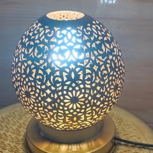 May include: A spherical lamp with a detailed, cut-out pattern. The lamp emits a warm glow, and sits on a round, gold-coloured base. The lamp's design is reminiscent of Moroccan or Middle Eastern decor.