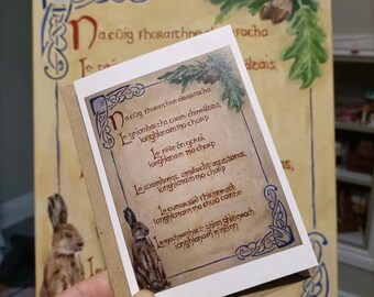 May include: A greeting card with a brown kraft paper backing, featuring a printed poem in Gaelic script. The card has a decorative border and an illustration of a hare. The background shows a larger painting with Gaelic text and a painted hare.